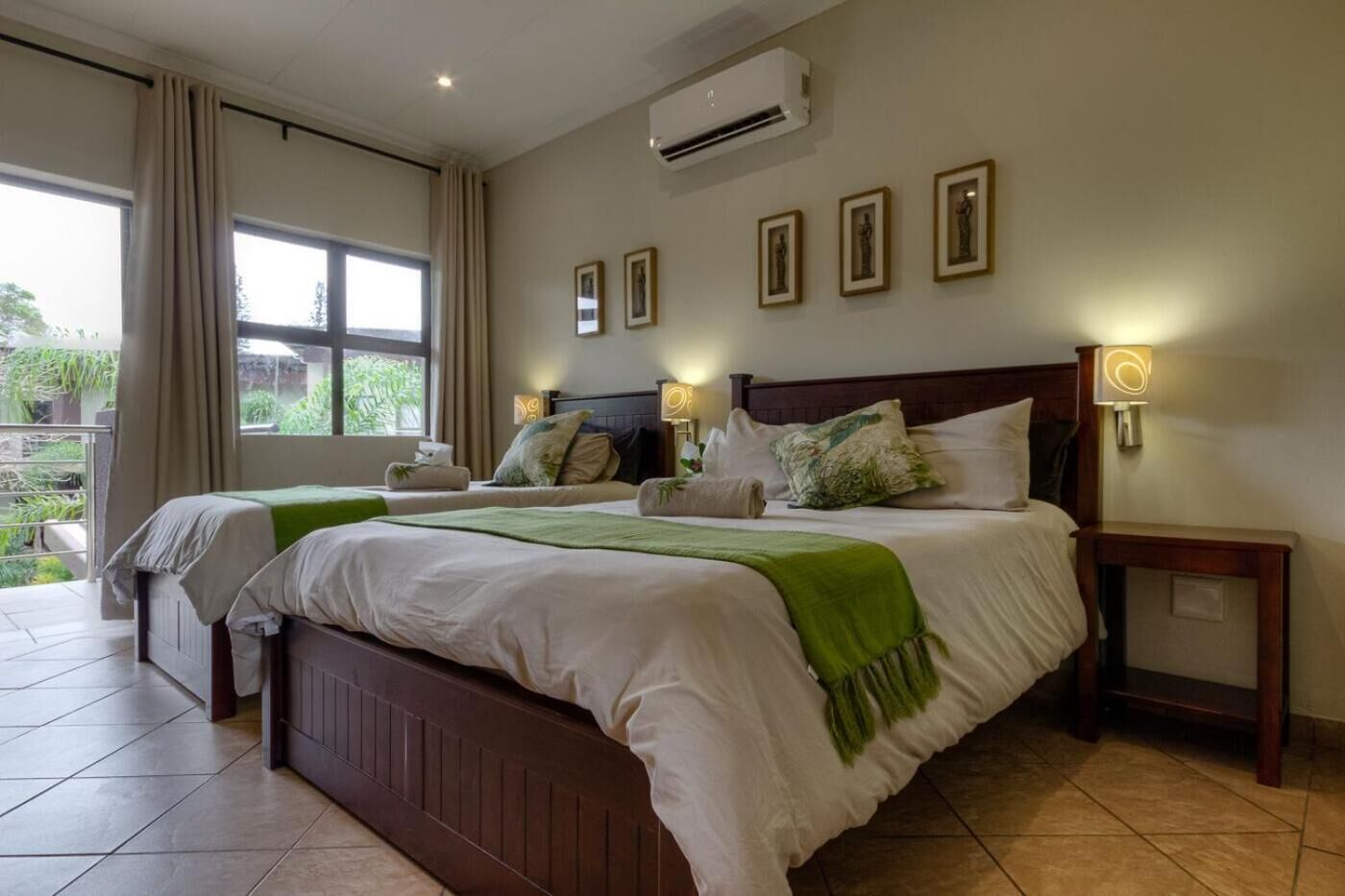 A hotel room with two beds, a large window with curtains, a wall-mounted air conditioner, and framed pictures.