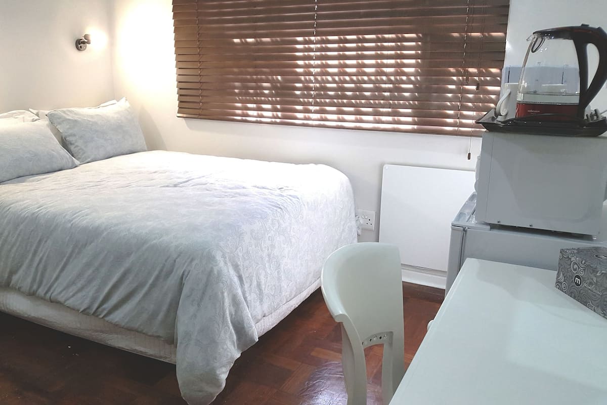 Une chambre à coucher comprenant un lit, une chaise, un bureau, un radiateur et une fenêtre avec des stores.
