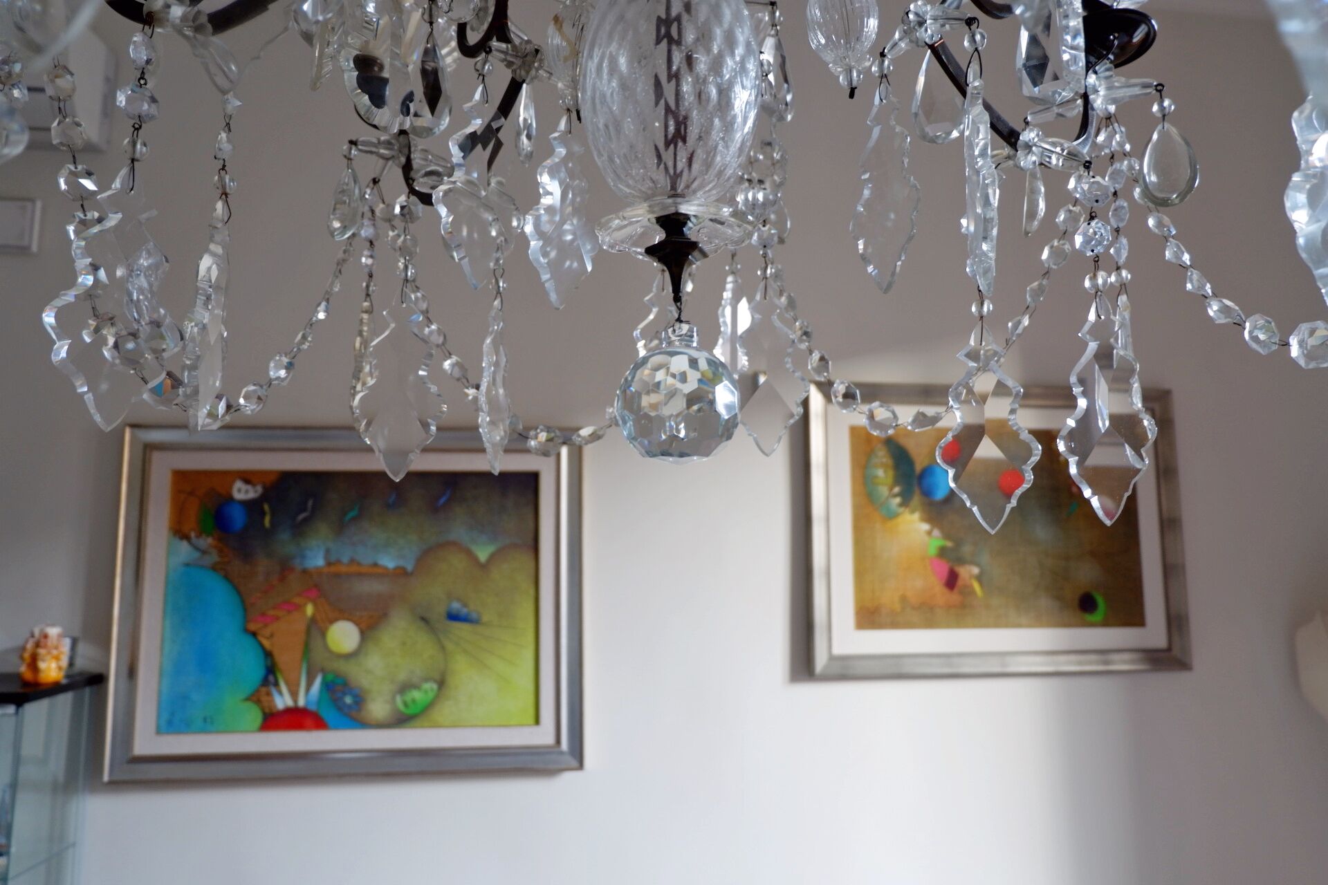 A room with a chandelier made of clear crystals and two framed paintings on the wall.