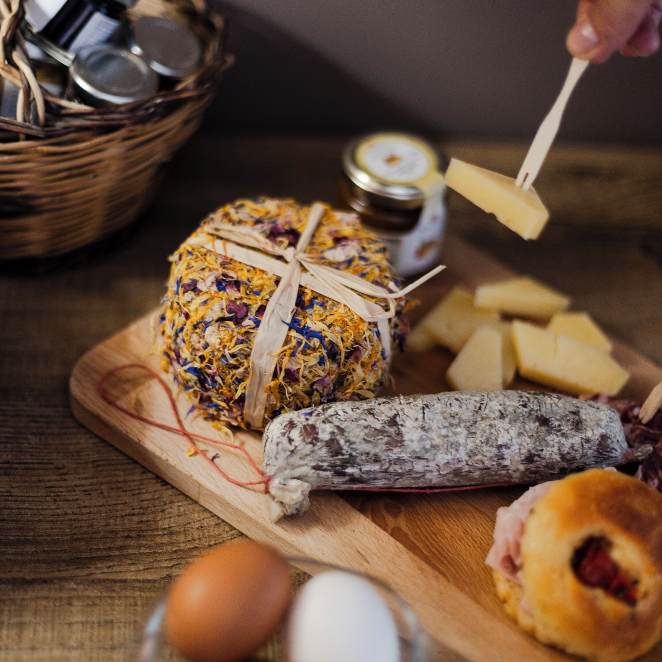 A wooden cutting board with a wrapped cheese, a cured sausage, and some cheese cubes.