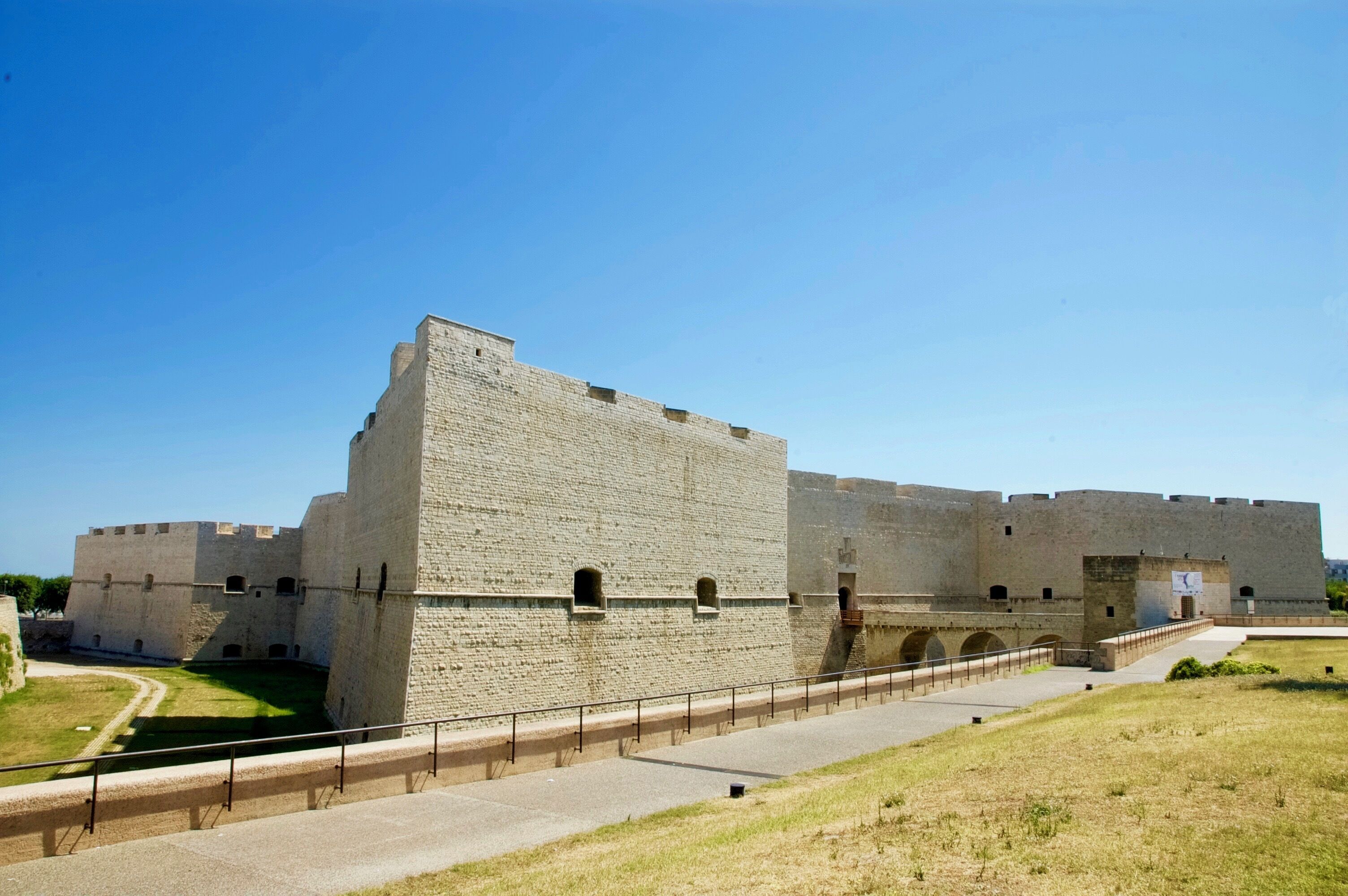 Un'imponente fortezza storica in pietra con un cortile centrale, un percorso lastricato e una zona verde antistante.