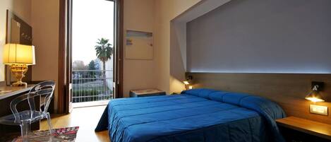 A hotel room with a large bed, a desk with a lamp, a chair, and a balcony with a view.