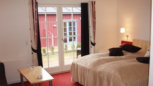 A hotel room with two beds, a small table, a lamp, and a view of a red building through the door. - Gerlev Kro & Hotel (Jægerspris)