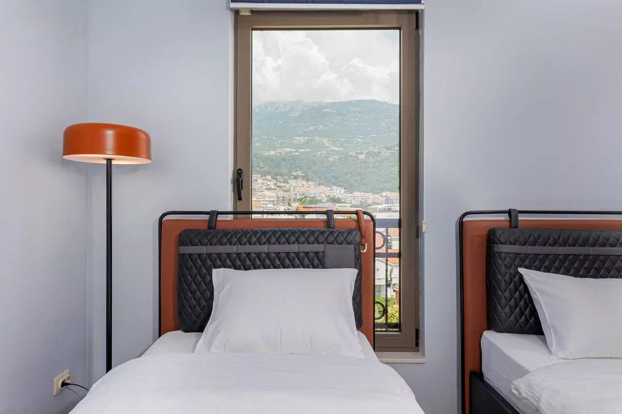 Habitación de hotel con dos camas, una ventana que ofrece vistas a una ciudad y montañas, y una lámpara de pie.