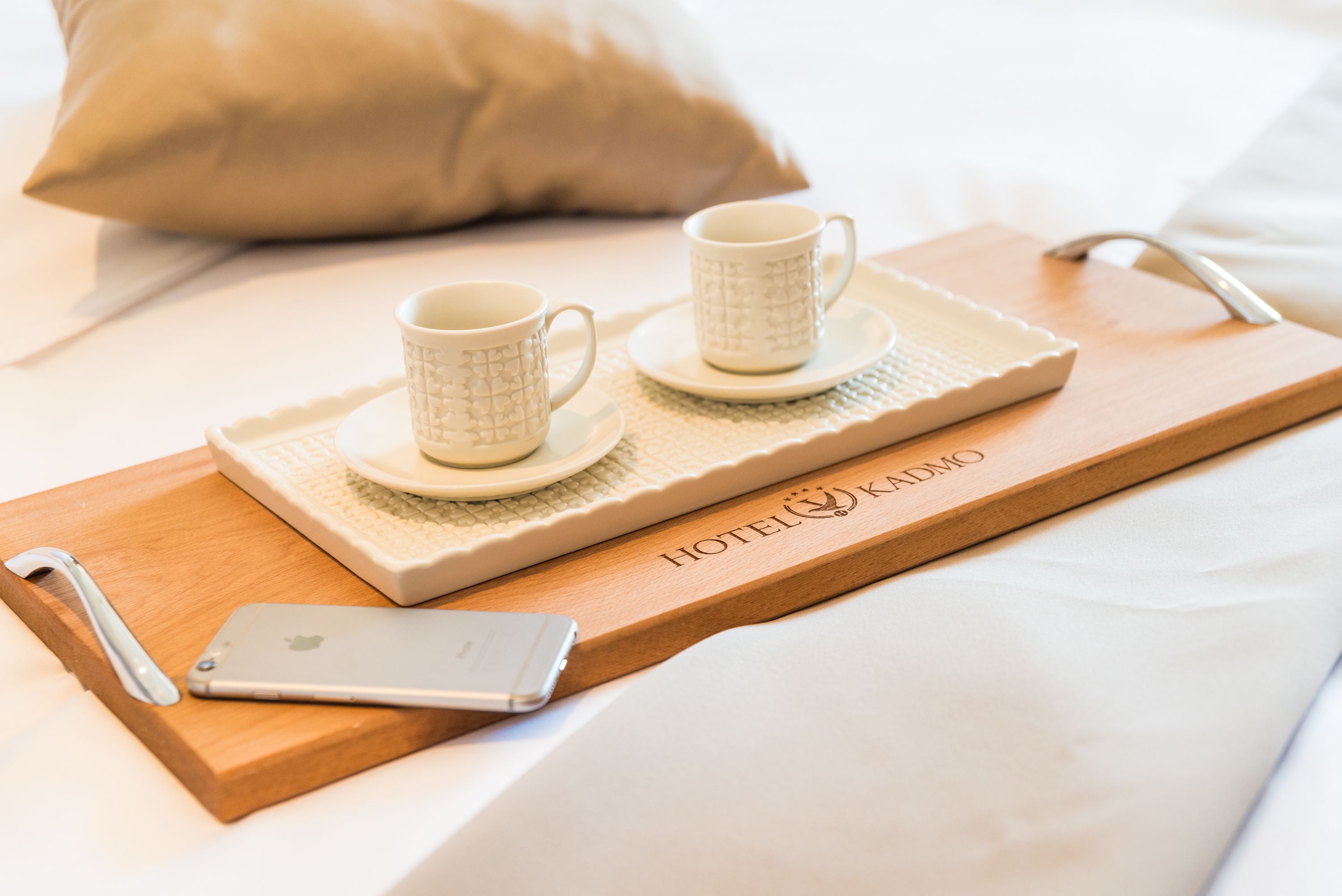 Una bandeja de madera con dos tazas y platillos blancos, un teléfono inteligente y una almohada sobre una cama.