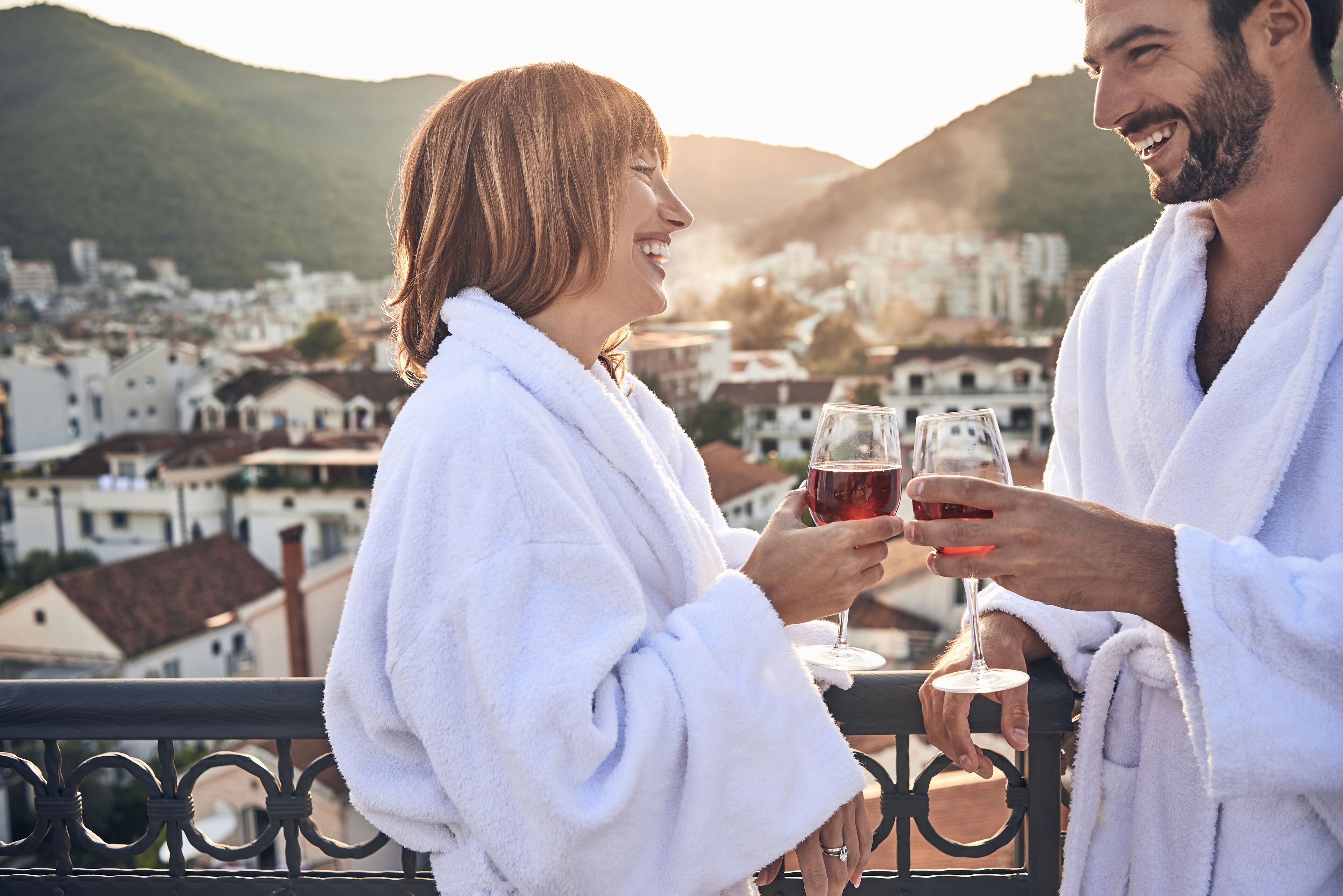 A couple in white bathrobes holding wine glasses, overlooking a town with buildings and hills in the background.