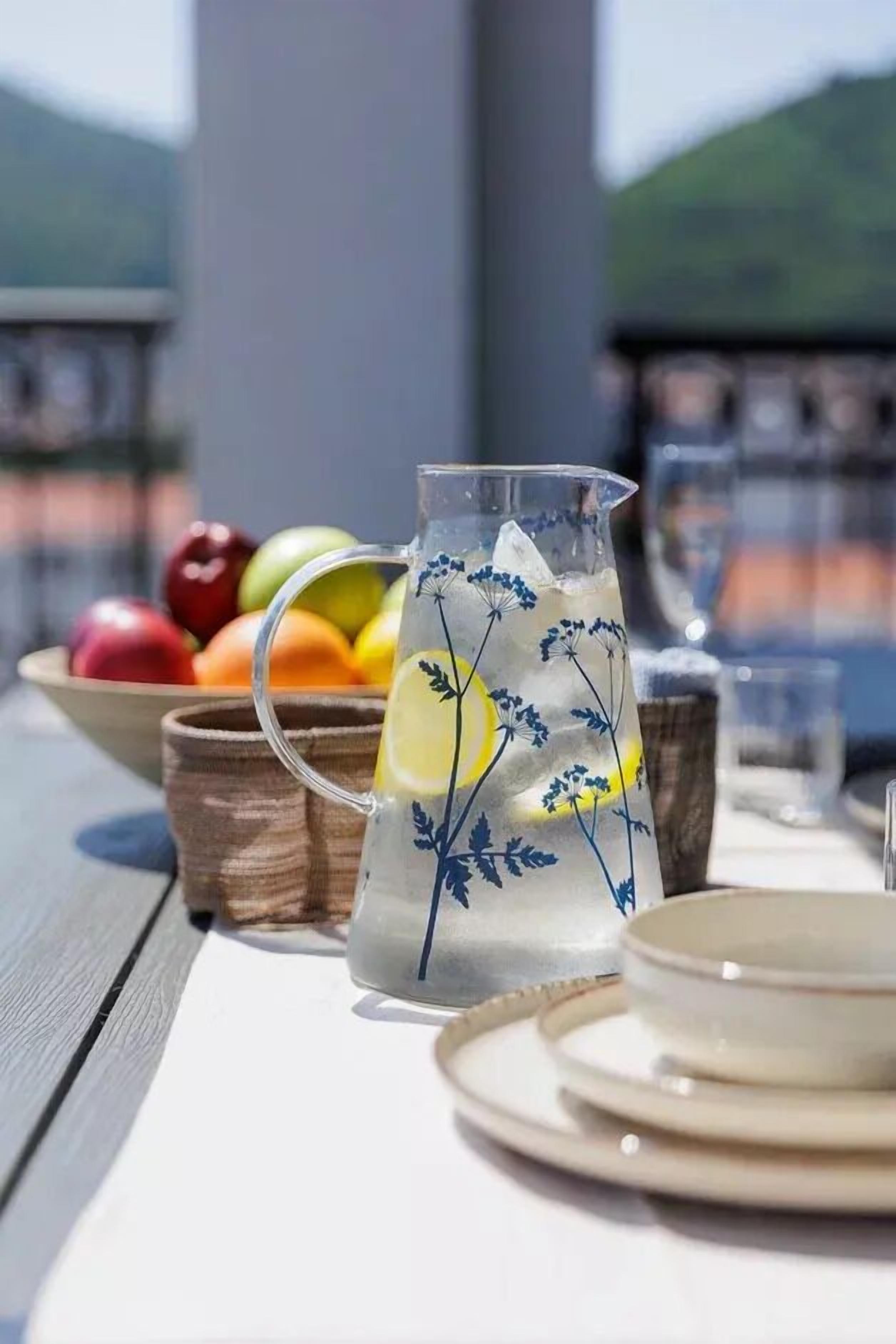 A clear glass pitcher with ice and lemon slices, placed on a table with a bowl of fruit in the background.