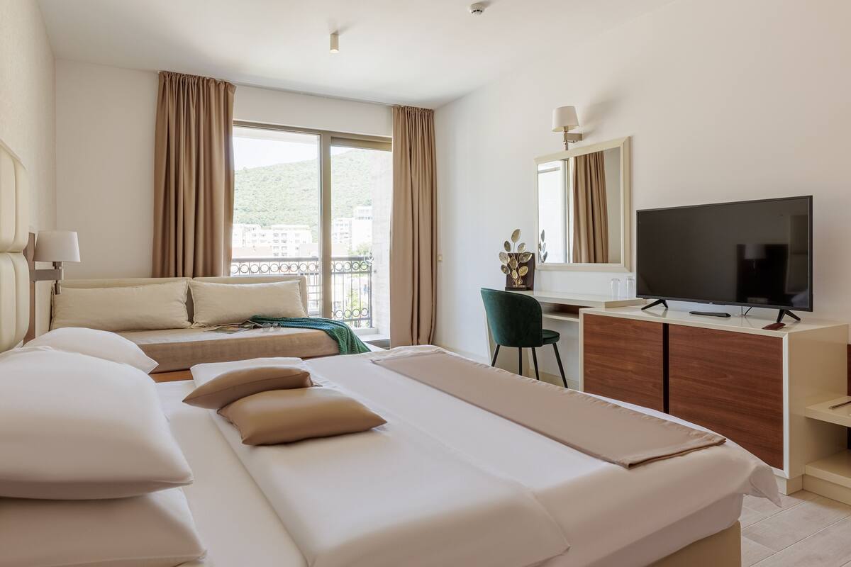 A hotel room with a large bed, a desk with a TV, and a balcony with curtains.