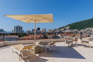 Una terraza en la azotea con una mesa, sillas, un paraguas grande y vistas a un paisaje urbano con edificios y una colina.