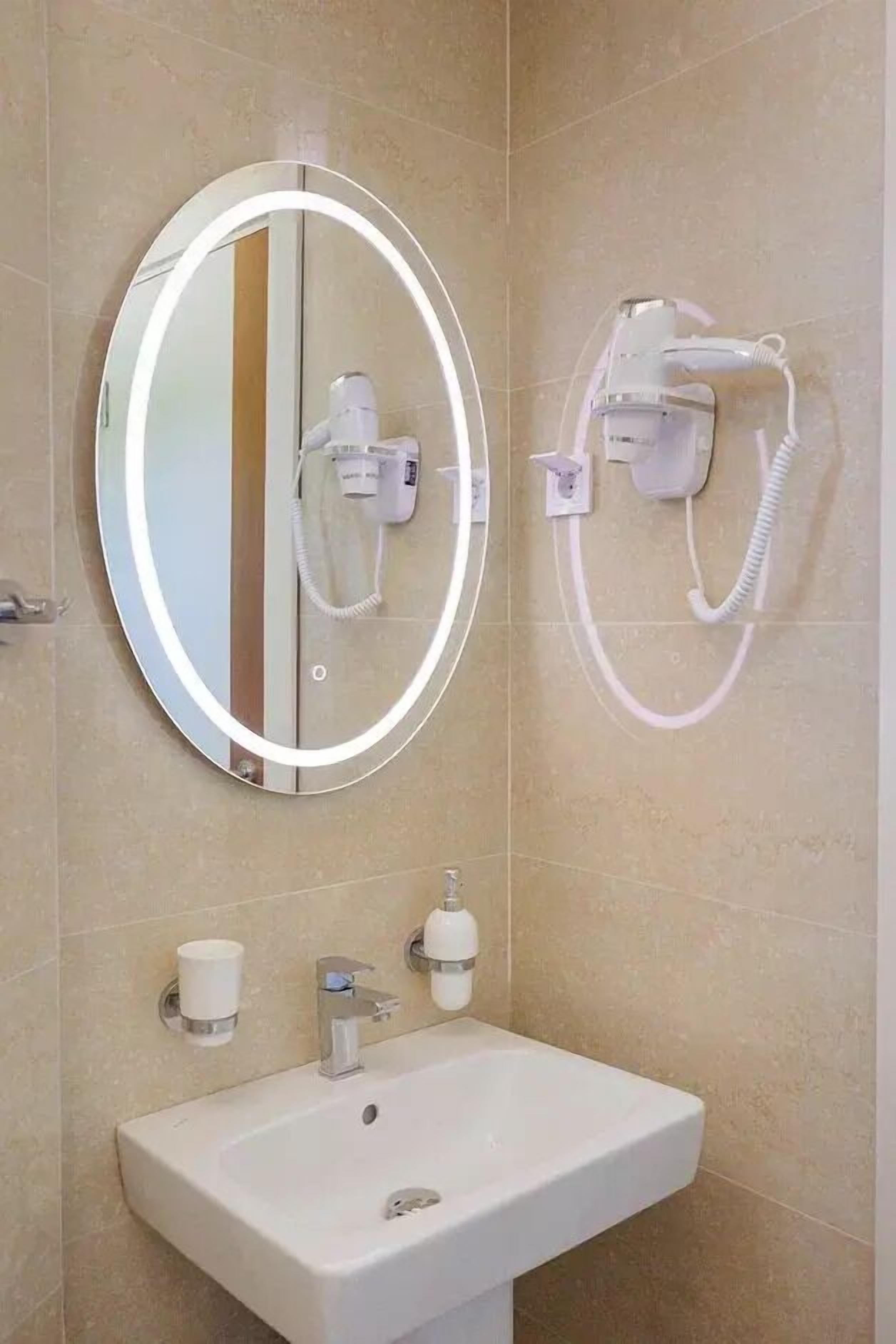 A bathroom with a round mirror, a hairdryer, a hand dryer, and a sink with soap dispensers.