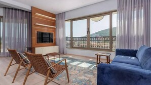 A modern living room with a blue sofa, wooden chairs, a TV, and a large window with a view of a hill. - Hotel Kadmo by Aycon (Budva)