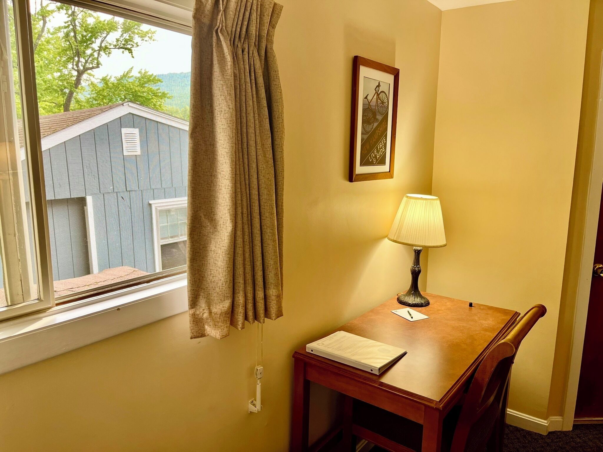 Une chambre d’angle avec une fenêtre, une lampe, un tableau encadré et une petite table sur laquelle se trouve un carnet.