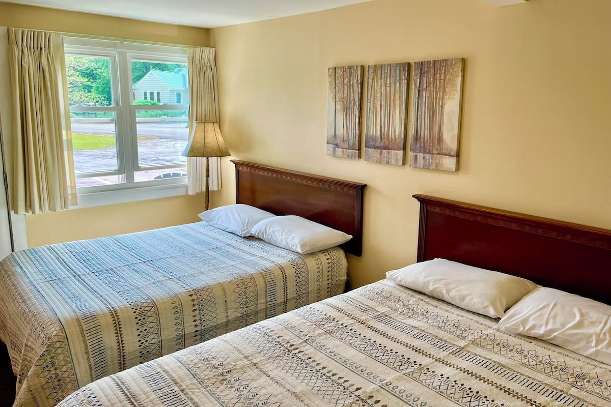 A hotel room with two beds, a window with curtains, a lamp, and three wall-mounted paintings.