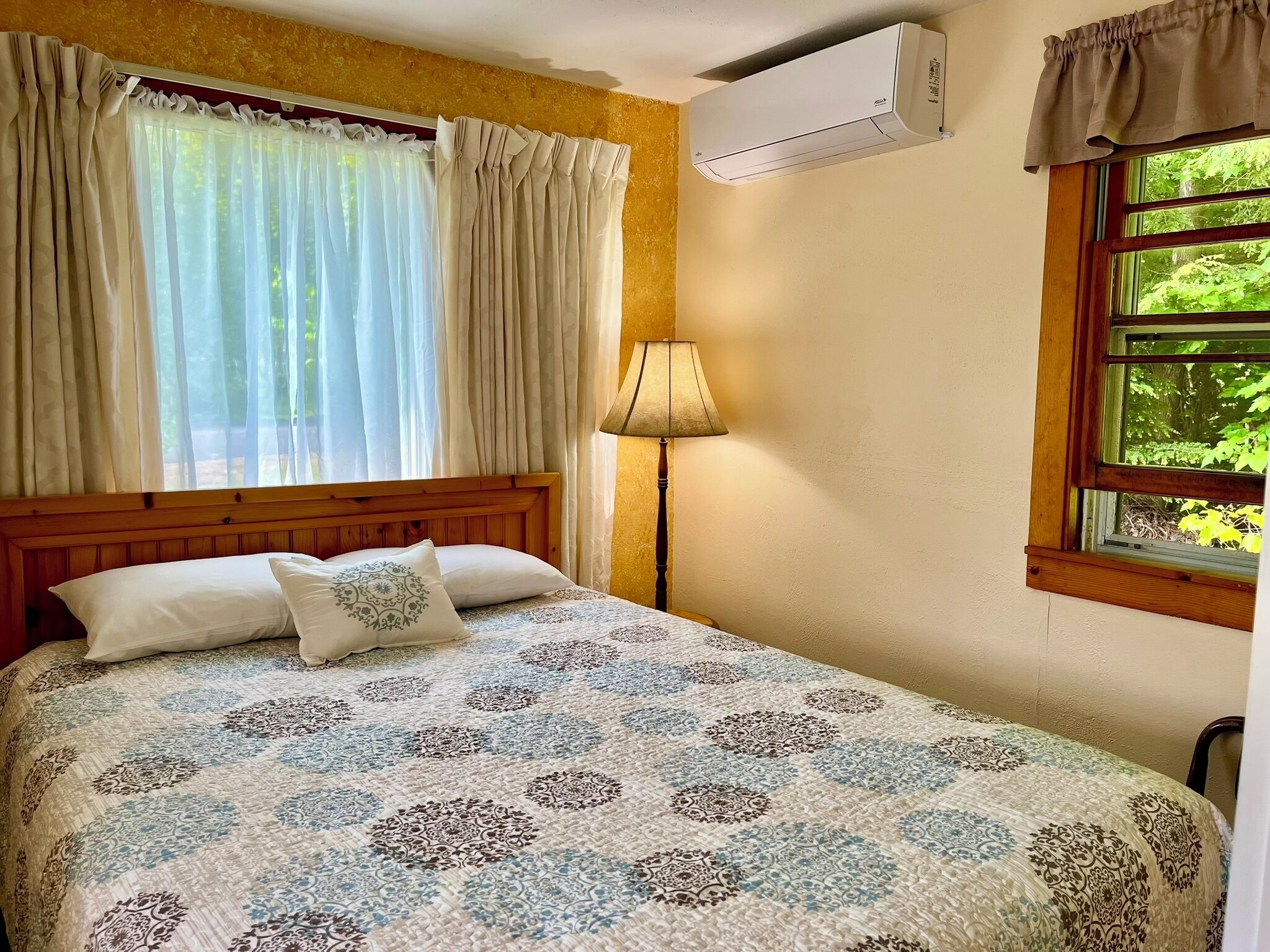 A bedroom with a bed, curtains, an air conditioner, a lamp, and a window with a view of greenery.