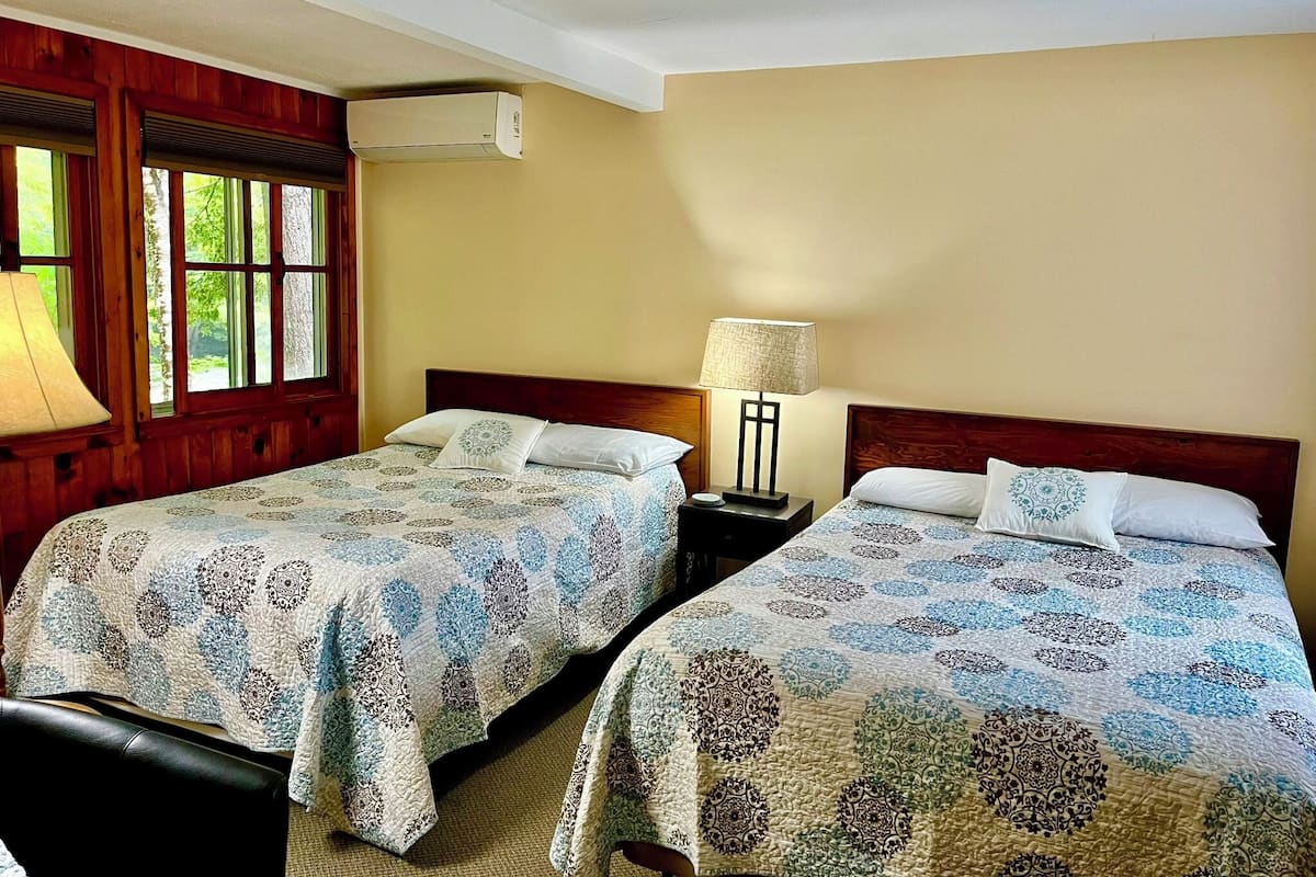A hotel room with two single beds, a wooden headboard, a nightstand with a lamp, and an air conditioning unit.