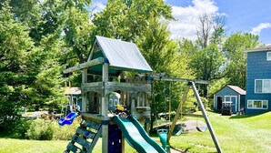 Una struttura di gioco in legno con scivolo e area dedicata all'arrampicata, immersa nel prato e tra gli alberi.