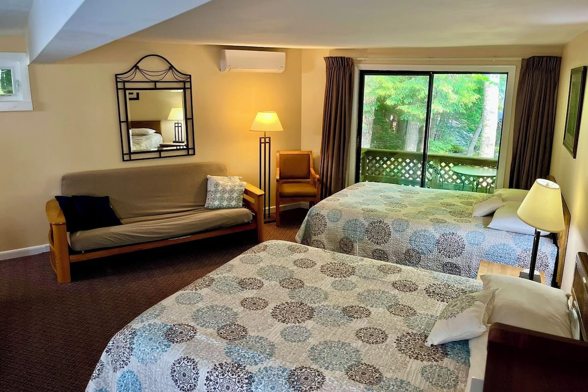 A hotel room with two beds, a sofa, a mirror, a lamp, and a window with a view of greenery.