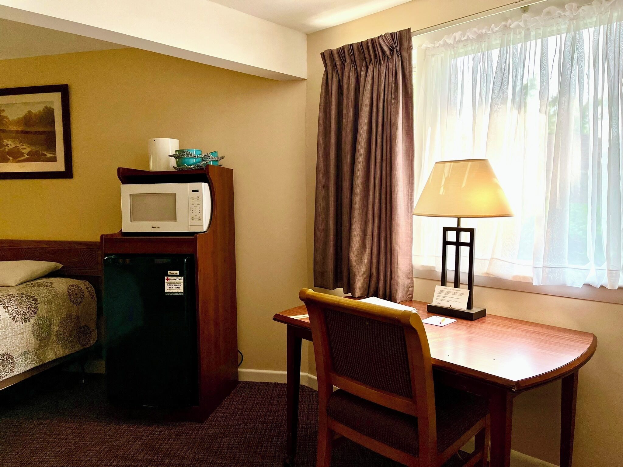 A hotel room with a bed, a desk with a chair, a microwave, a refrigerator, a lamp, and a window with curtains.