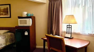 A hotel room with a bed, a desk with a chair, a microwave, a refrigerator, a lamp, and a window with curtains.