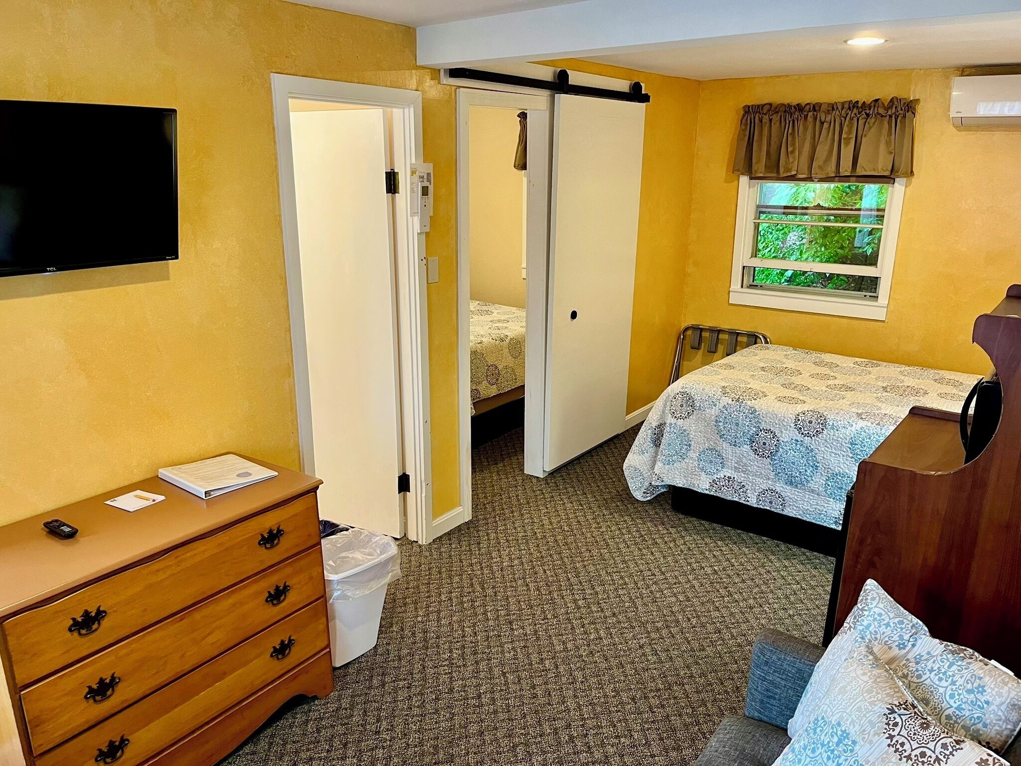 A hotel room with a bed, a dresser, a TV, and a window with a view of greenery.
