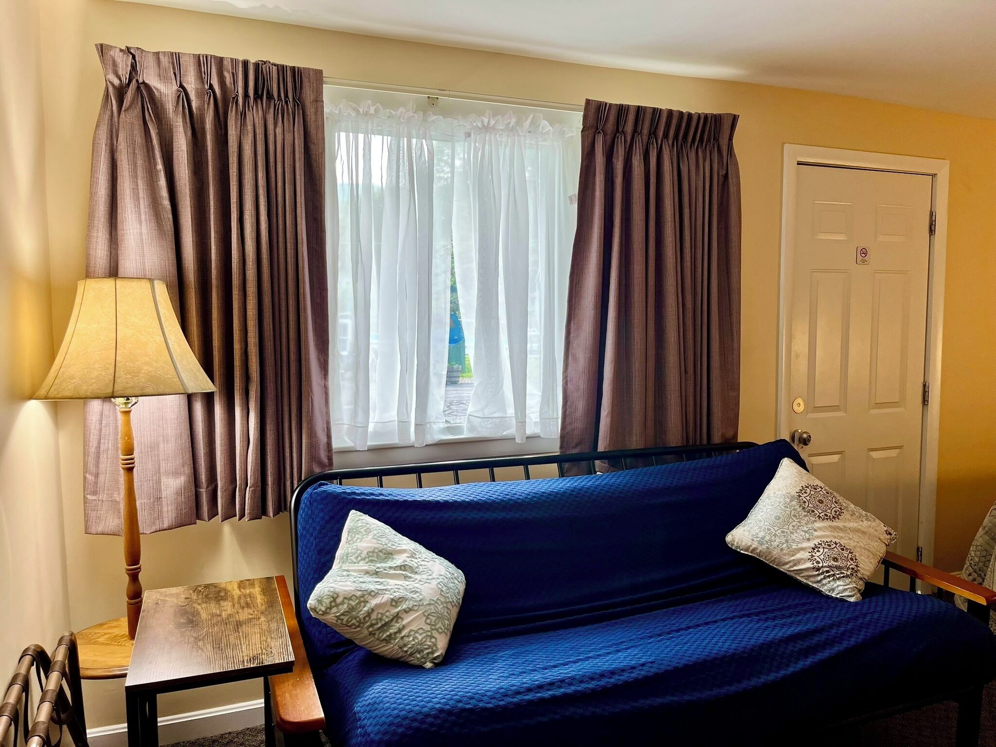 Una habitación con sofá azul, dos almohadas, una lámpara, una silla y una ventana con cortinas.