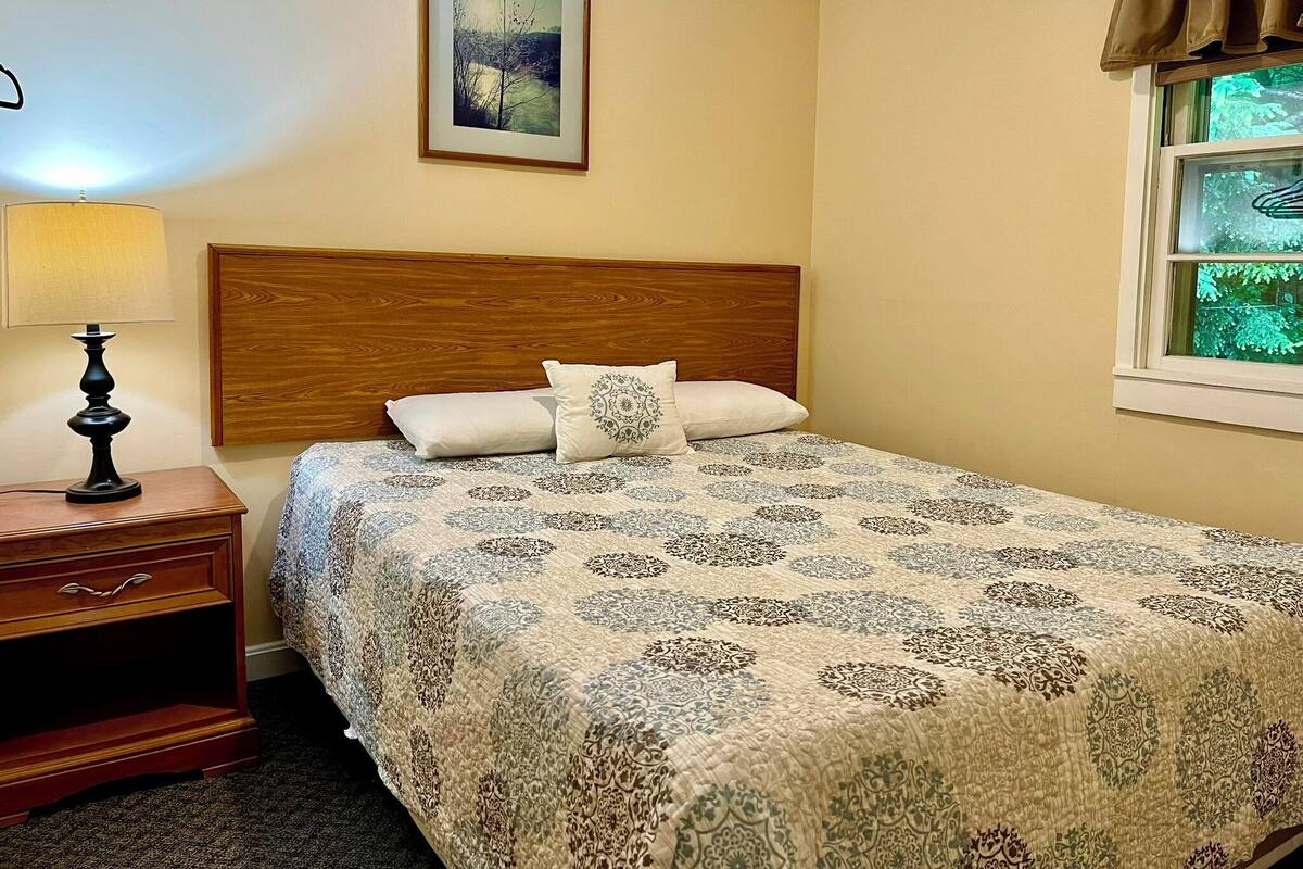 A neatly made bed with a patterned bedspread, a wooden headboard, a bedside table with a lamp, and a framed picture on the wall.