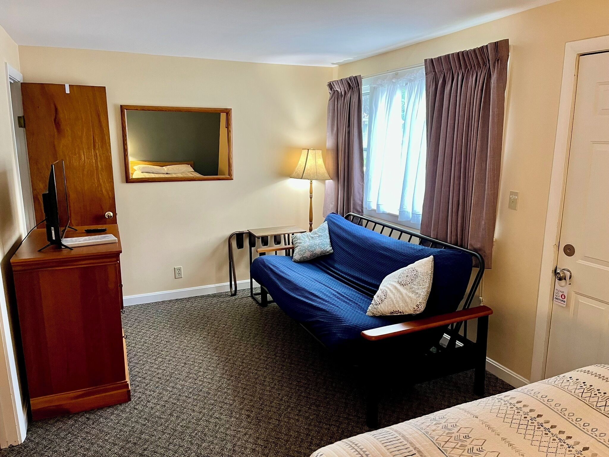 A compact hotel room with a single bed, a desk, a chair, a TV on a stand, a mirror, and a door with a sign.
