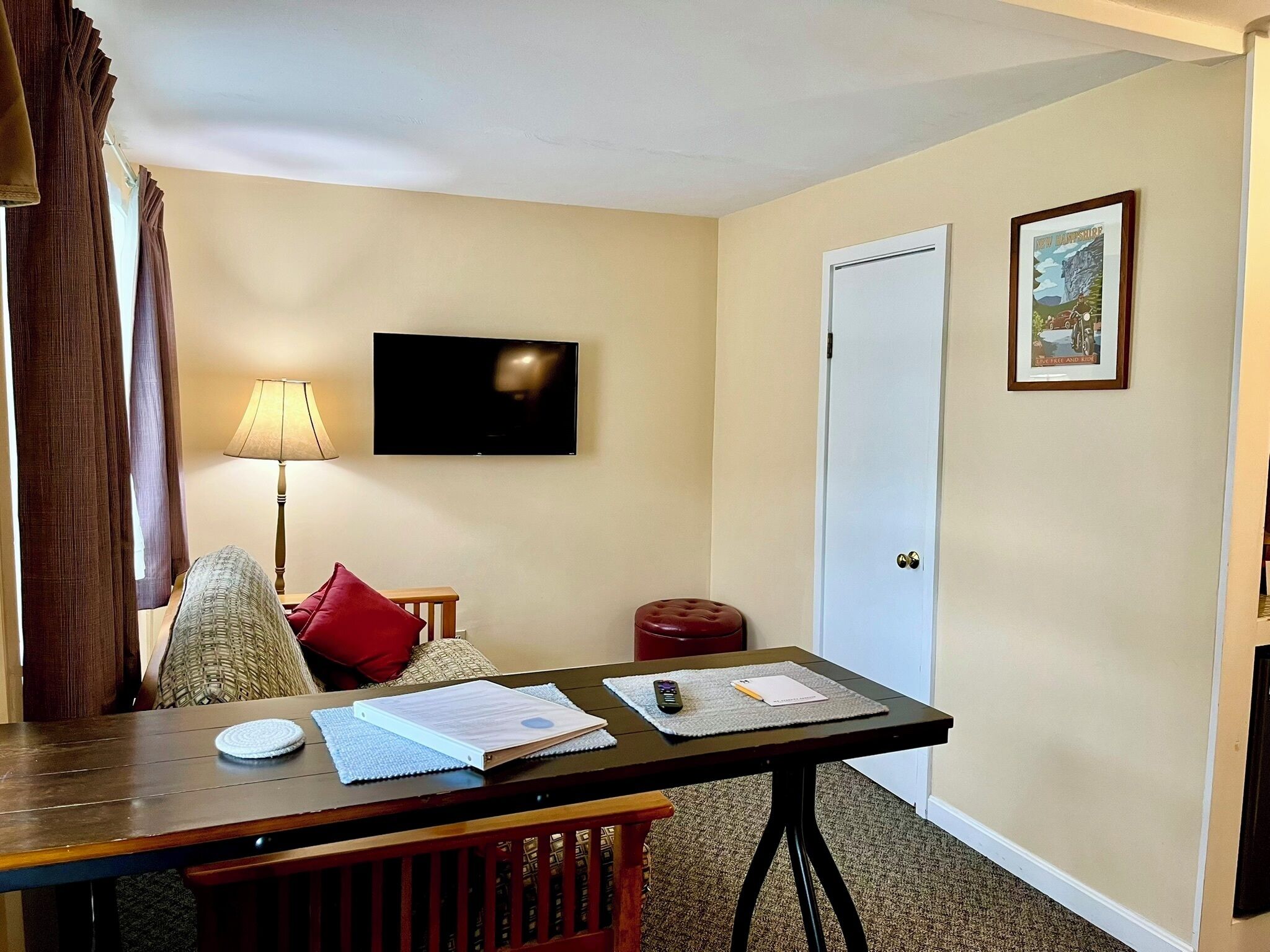 A hotel room with a wooden table, a television mounted on the wall, a door, and a framed picture on the wall.