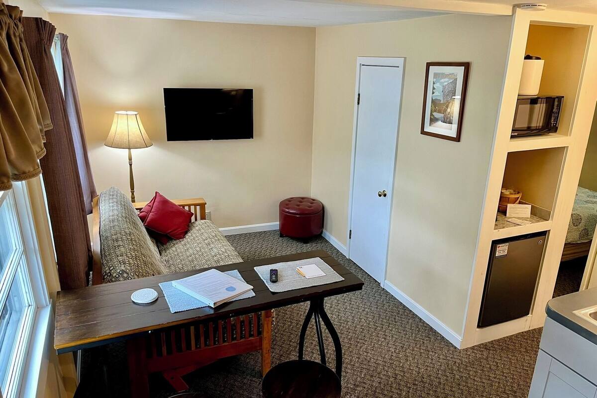 A compact hotel room with a sofa, a small table with papers, a lamp, a TV, a door, and a kitchenette.