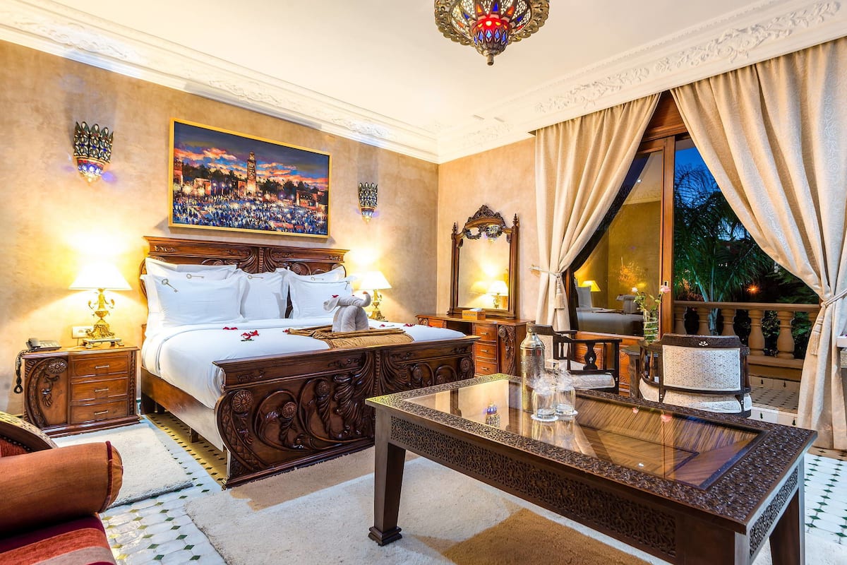 A luxurious hotel room with a large bed, a glass-top table, a painting of a cityscape, and a view of the outdoors.