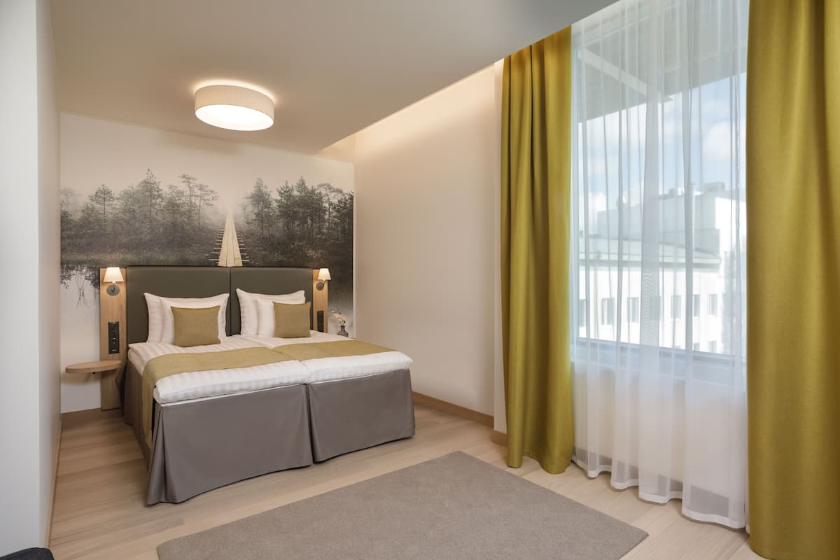 A modern hotel room with a bed, bedside tables, a nightstand, a large window with sheer curtains, and a wall-mounted light fixture.
