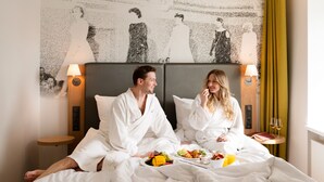 Um casal em quarto de hotel, com cama, bandeja de café da manhã e parede com mural em preto e branco.