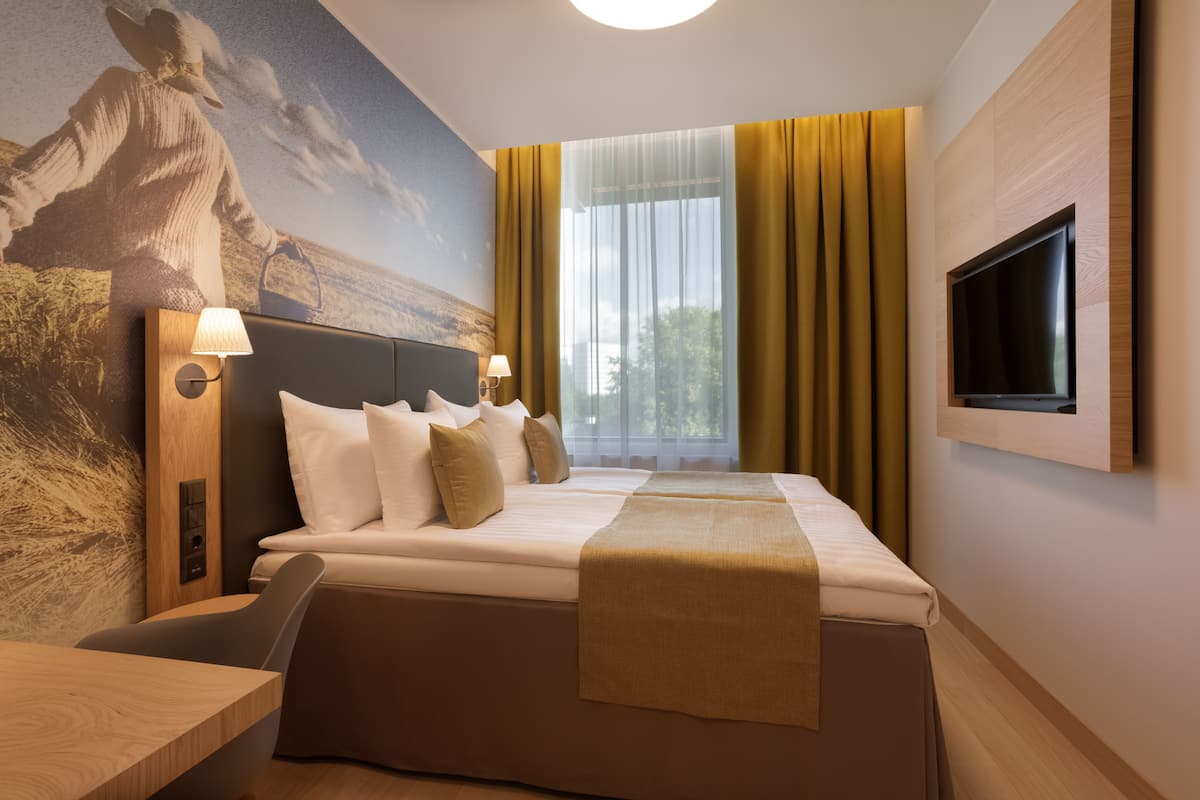 A hotel room with a large bed, a flat-screen TV mounted on the wall, a desk with a lamp, and a mural on the wall.
