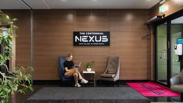 Un hall d’hôtel moderne avec une grande enseigne indiquant « The Centennial Nexus Hotel Tallinn ». On y trouve deux espaces détente avec des chaises et une petite table agrémentée d’une plante.