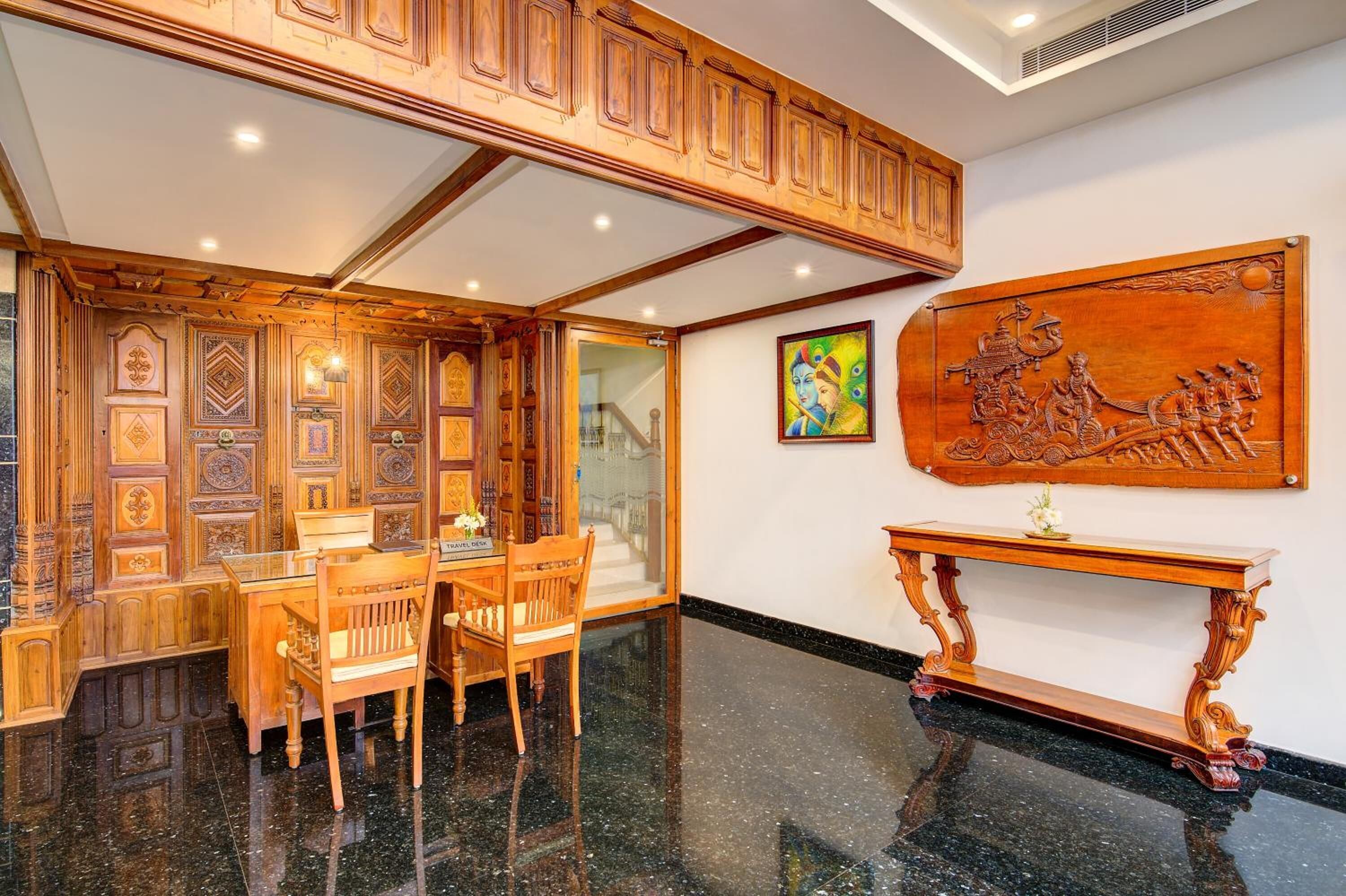 A hotel lobby with a wooden reception desk, a dining area with chairs, a staircase, and a large carved wooden artwork on the wall.