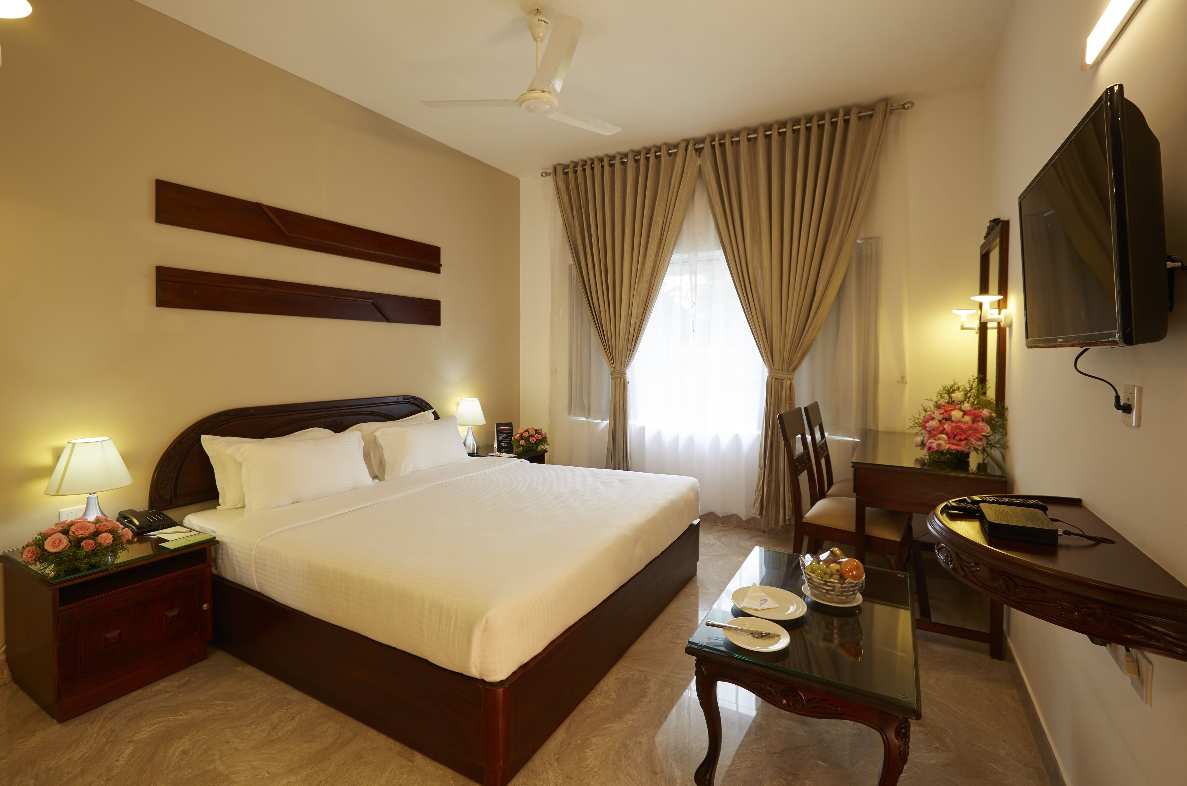 A hotel room with a large bed, bedside table with a lamp, a desk with a chair, a television, and a window with curtains.