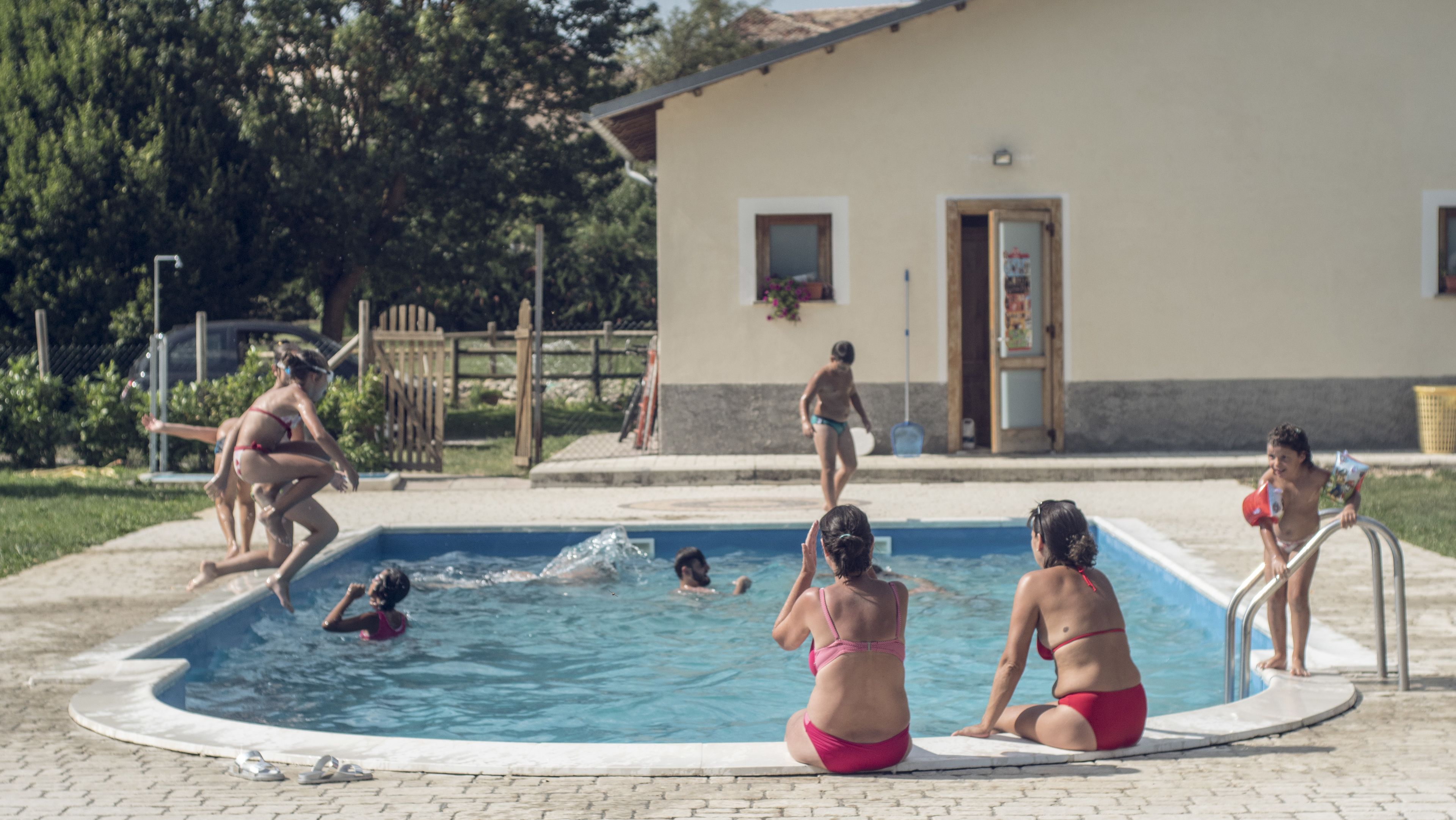 A swimming pool with people in and around it, a building with a door and windows, and a fence in the background.