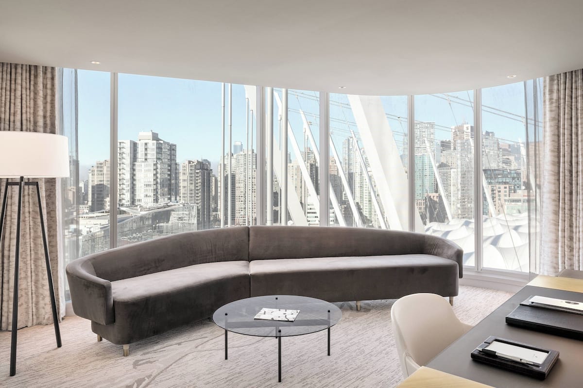 Una sala moderna con un sofá gris curvo, una mesa de centro redonda de vidrio y vistas a un paisaje urbano a través de ventanas amplias.