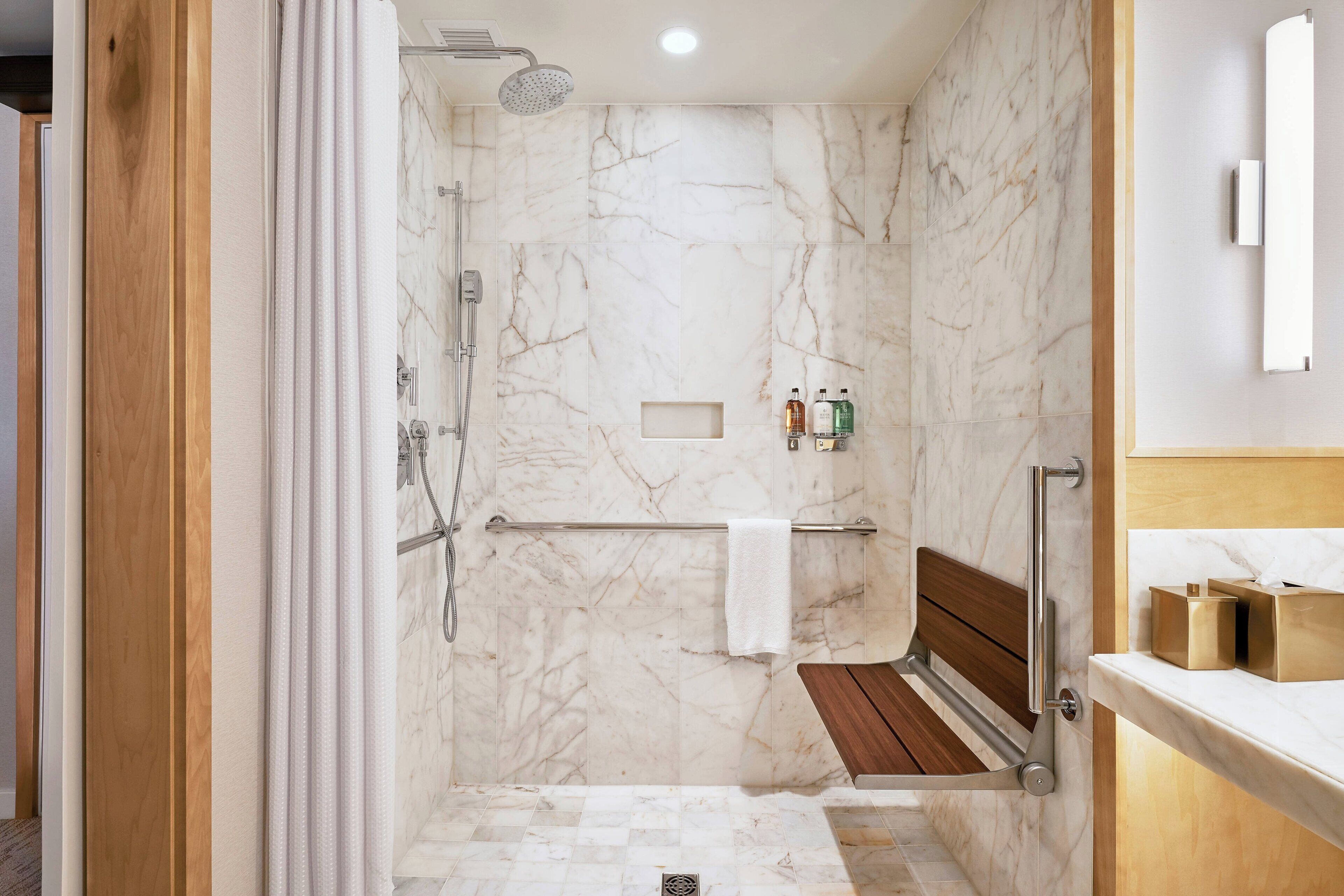 Un baño moderno con ducha, un banco de madera, paredes de mármol y un tocador con artículos de aseo.