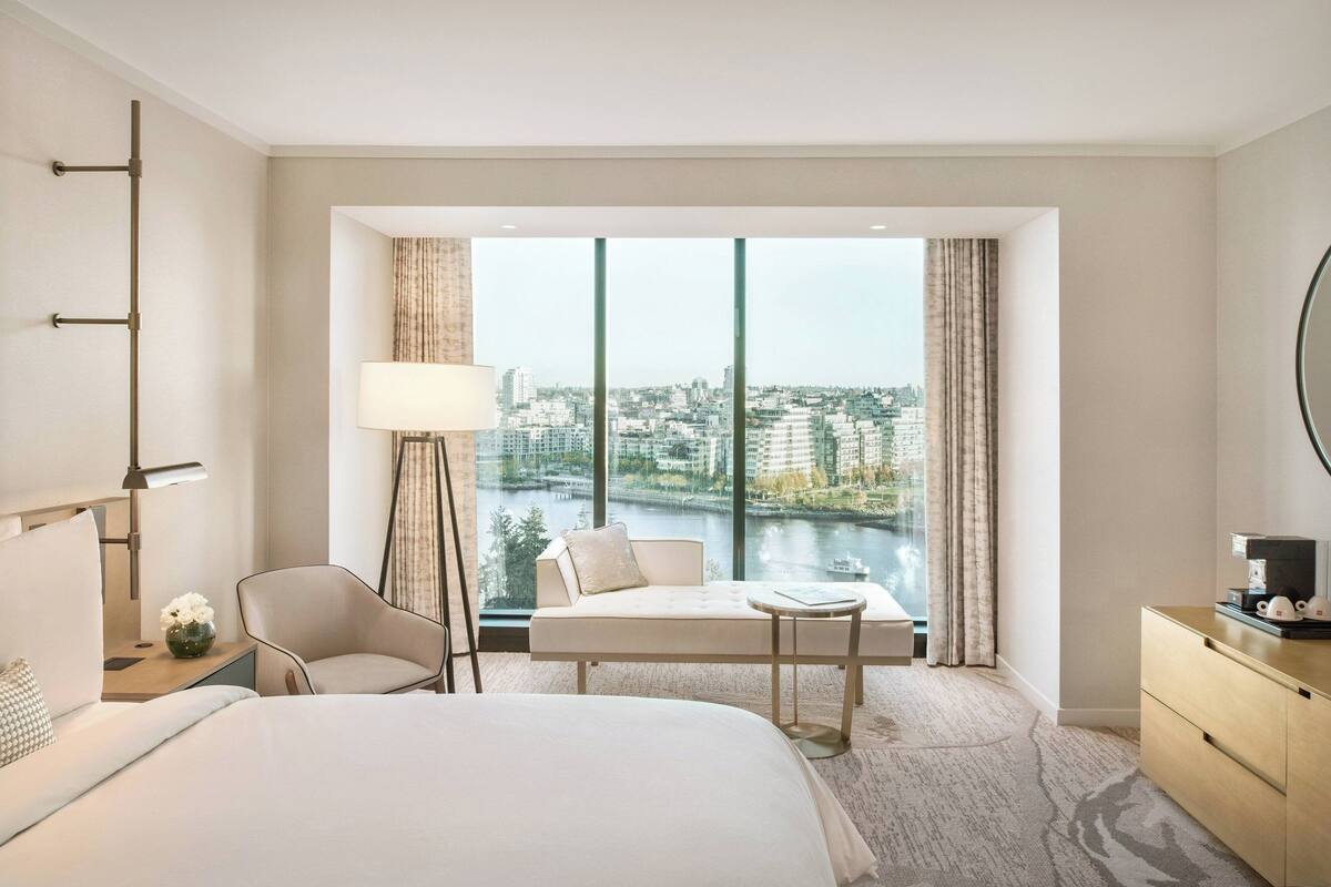 Una habitación de hotel moderna con una cama grande, una zona de estar con vistas a un paisaje urbano y un río, y un escritorio con una lámpara y una cafetera.