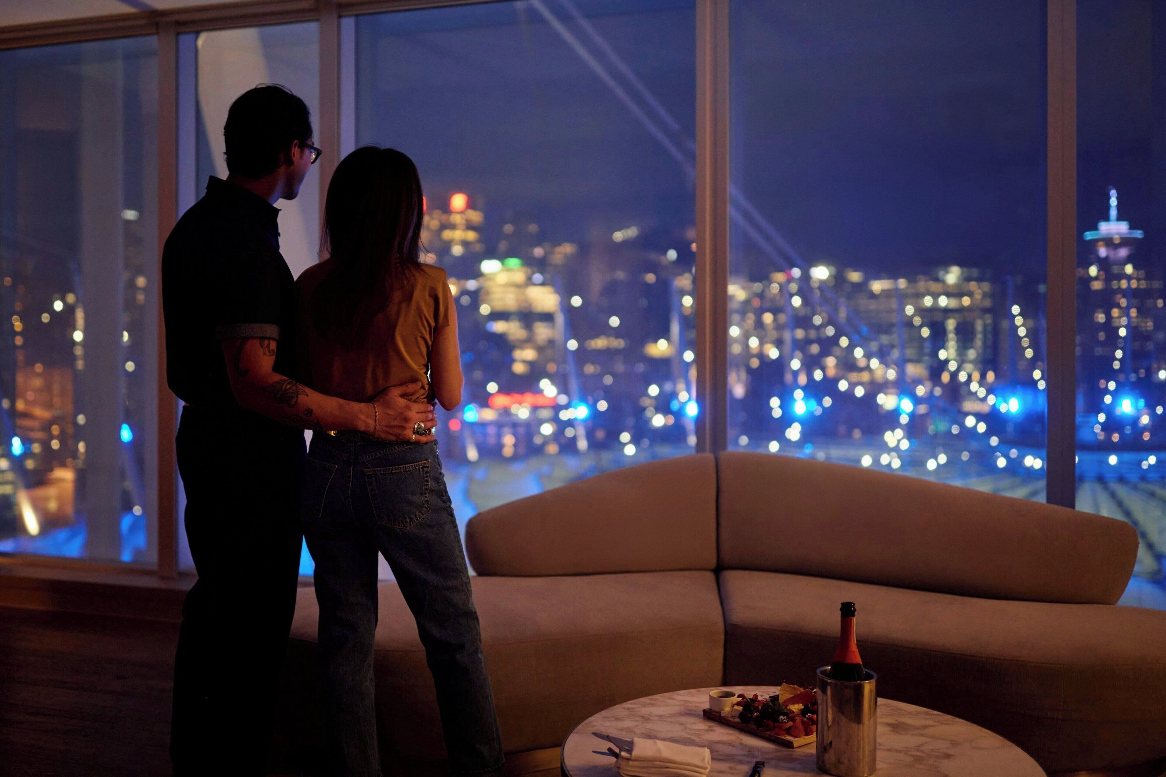 Una pareja de pie junto a un amplio ventanal con vista a un paisaje urbano nocturno.