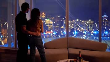 Una pareja de pie junto a un amplio ventanal con vista a un paisaje urbano nocturno.