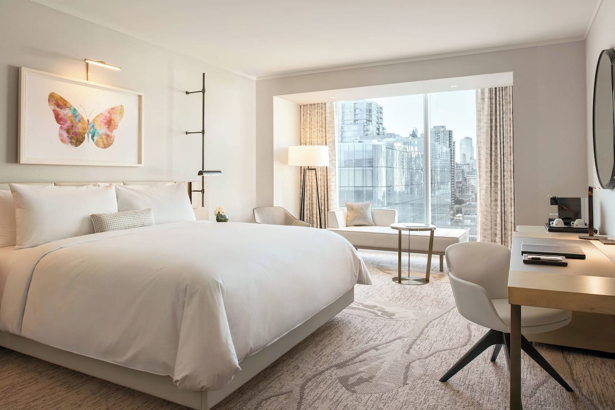 Una habitación de hotel moderna con una cama grande, un escritorio, una silla, un sofá y vistas de la ciudad a través de amplias ventanas.