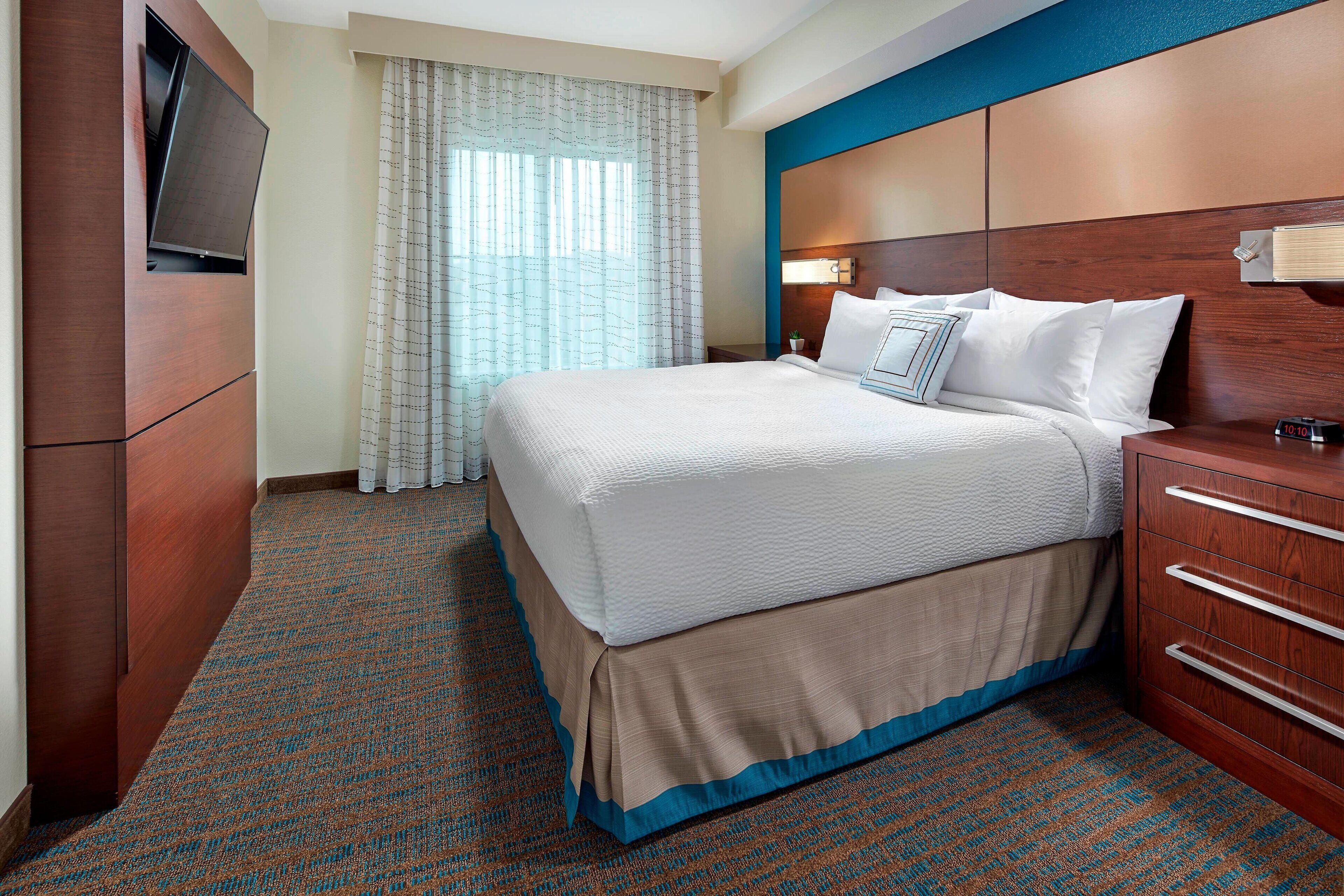 A hotel room with a large bed, a TV mounted on the wall, a bedside table with a lamp, and a window with curtains.