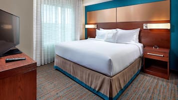 A hotel room with a large bed, a TV on a wooden desk, a nightstand with a lamp, and a window with sheer curtains.