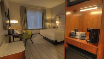 A hotel room with a bed, a desk with a lamp, a chair, a refrigerator, and a coffee maker.