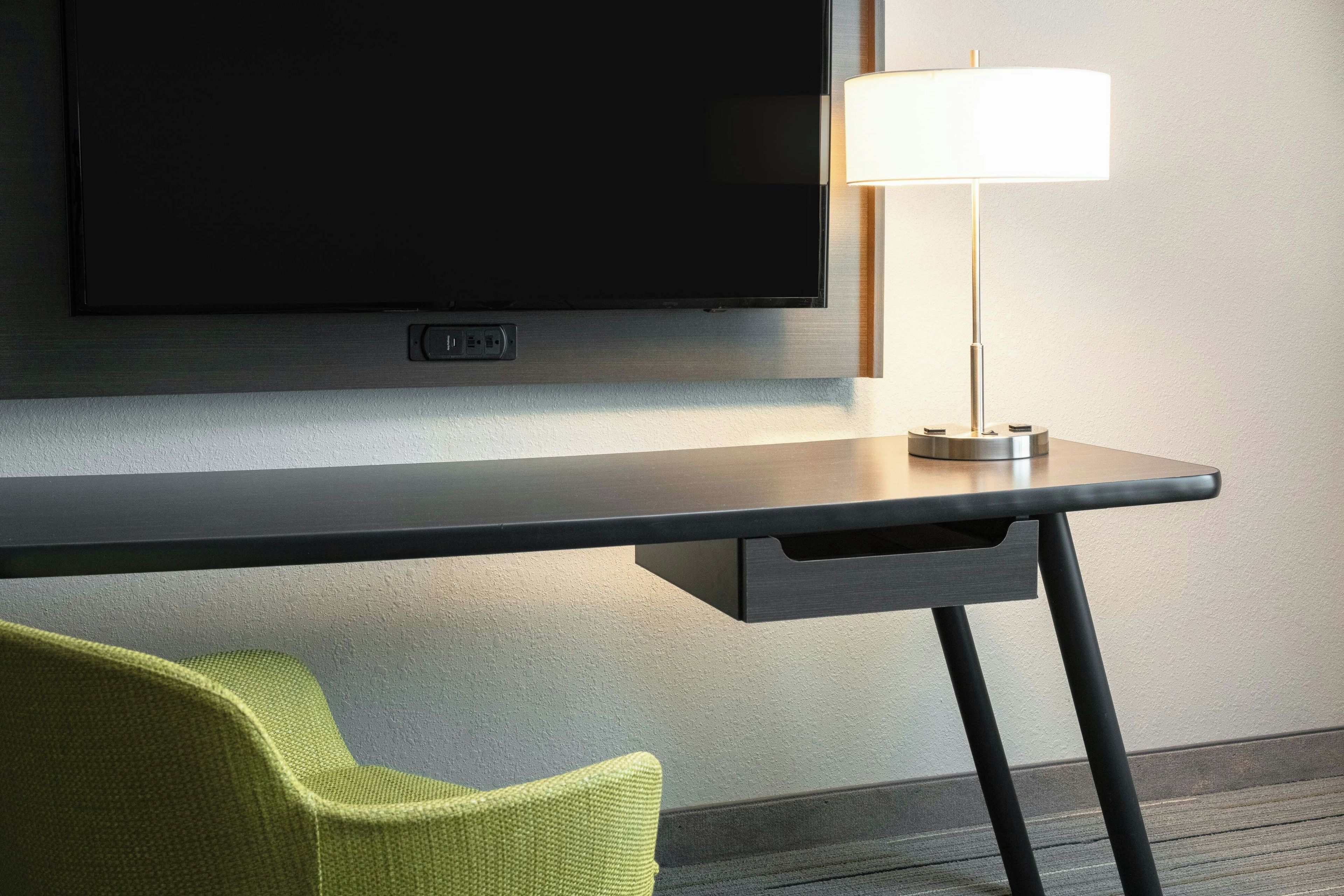A modern hotel room with a flat-screen TV mounted on the wall, a desk with a lamp, and a green chair.