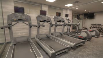 A row of treadmills with digital screens, a wall-mounted TV, and a rack of dumbbells.