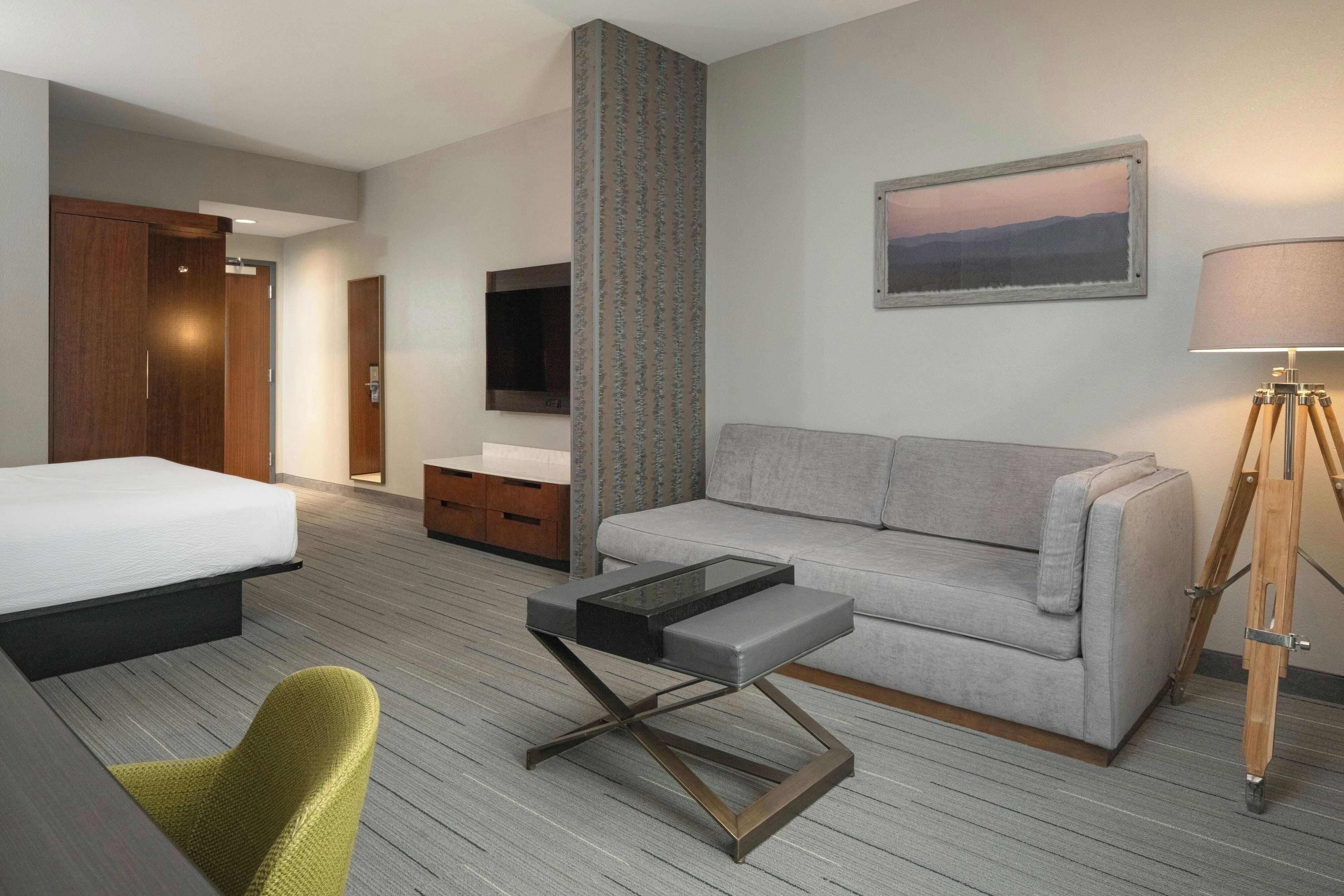 Quarto de hotel com cama, sofá, mesa pequena, cadeira, televisão, luminária e pintura na parede.