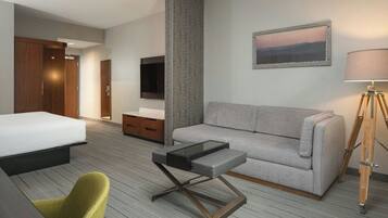 Quarto de hotel com cama, sofá, mesa pequena, cadeira, televisão, luminária e pintura na parede.
